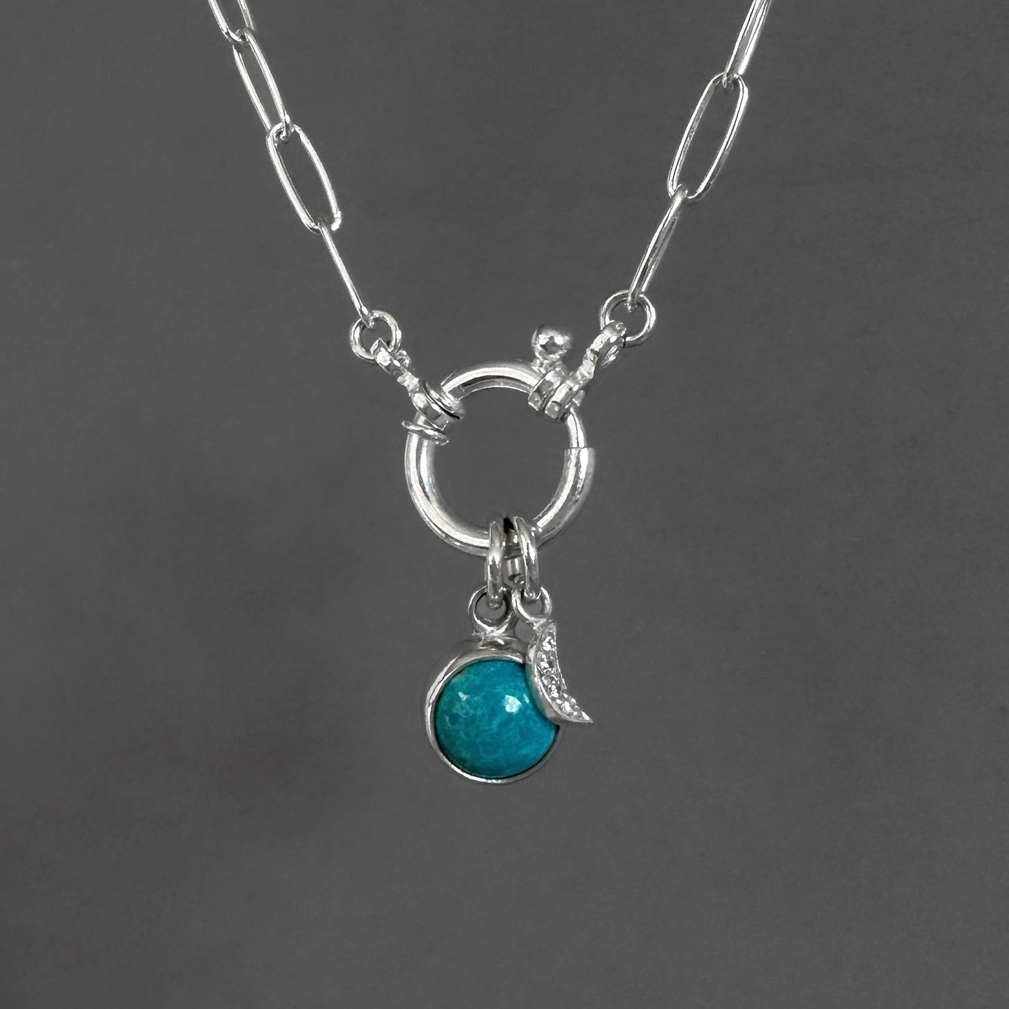 A silver paperclip chain necklace with a toggle clasp, featuring a circular ring and a dangling charm set with a round turquoise-blue stone and a small crescent moon, shown against a dark gray background.