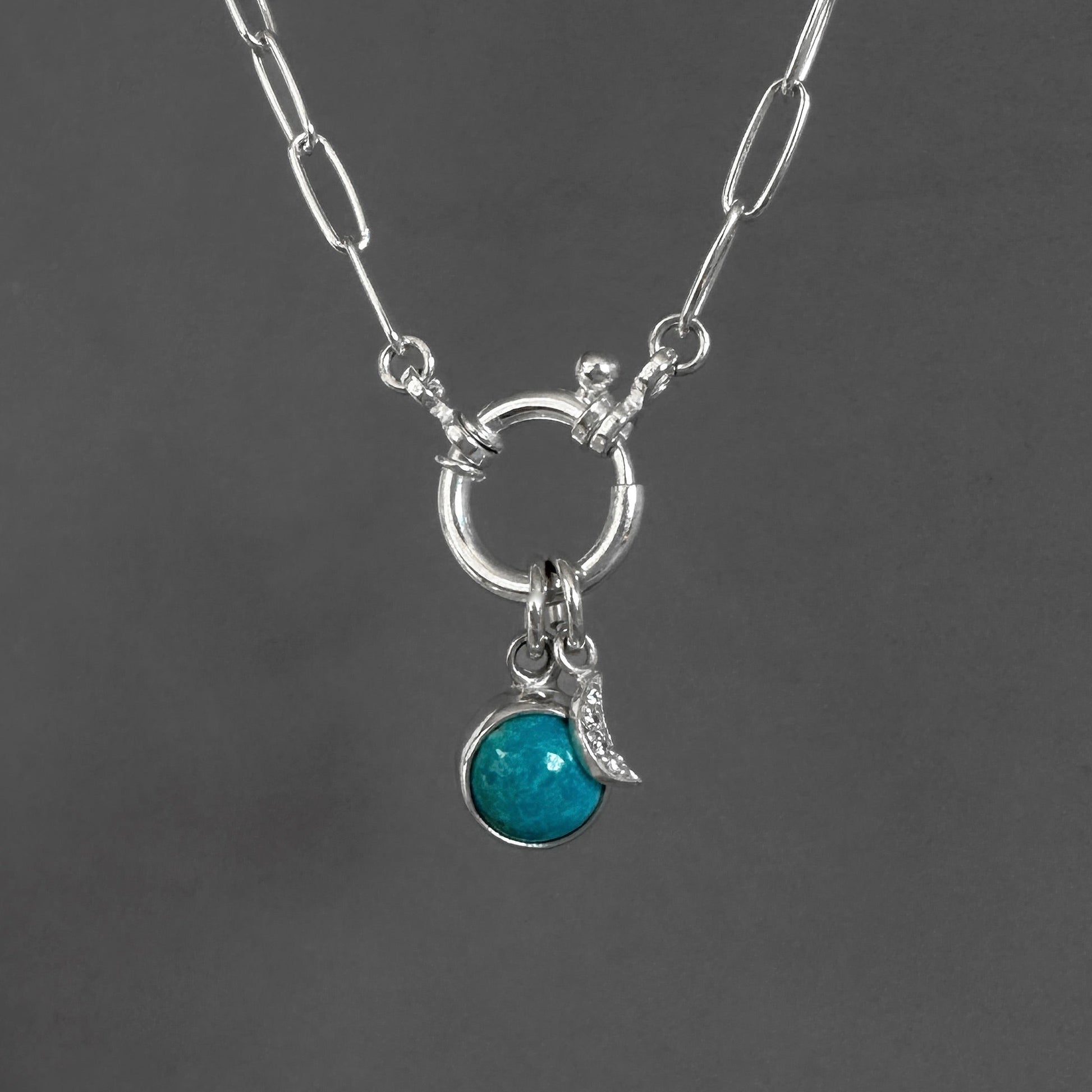 A silver paperclip chain necklace with a toggle clasp, featuring a circular ring and a dangling charm set with a round turquoise-blue stone and a small crescent moon, shown against a dark gray background.