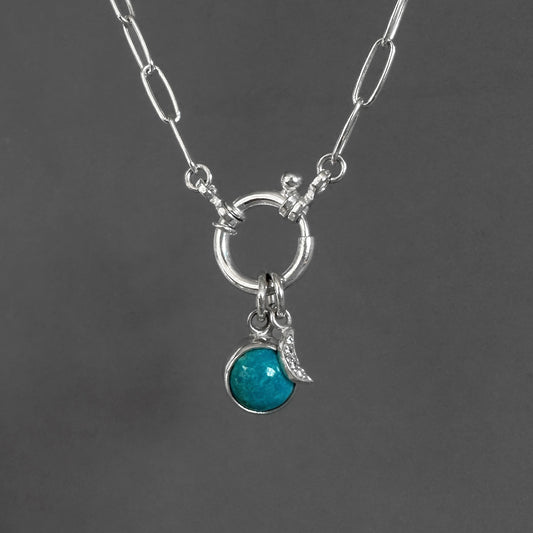 A silver paperclip chain necklace with a toggle clasp, featuring a circular ring and a dangling charm set with a round turquoise-blue stone and a small crescent moon, shown against a dark gray background.