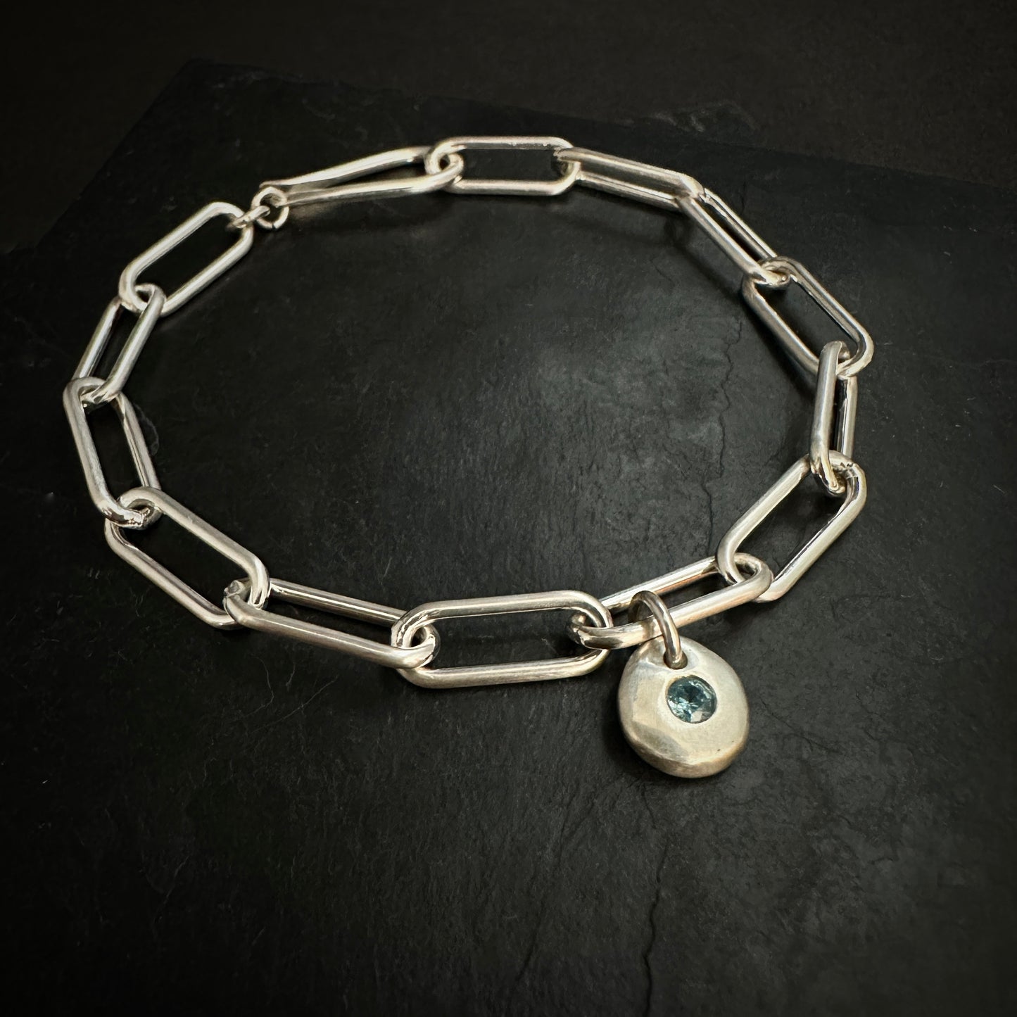 Silver chain bracelet with a heart-shaped charm featuring a blue gemstone on a black background