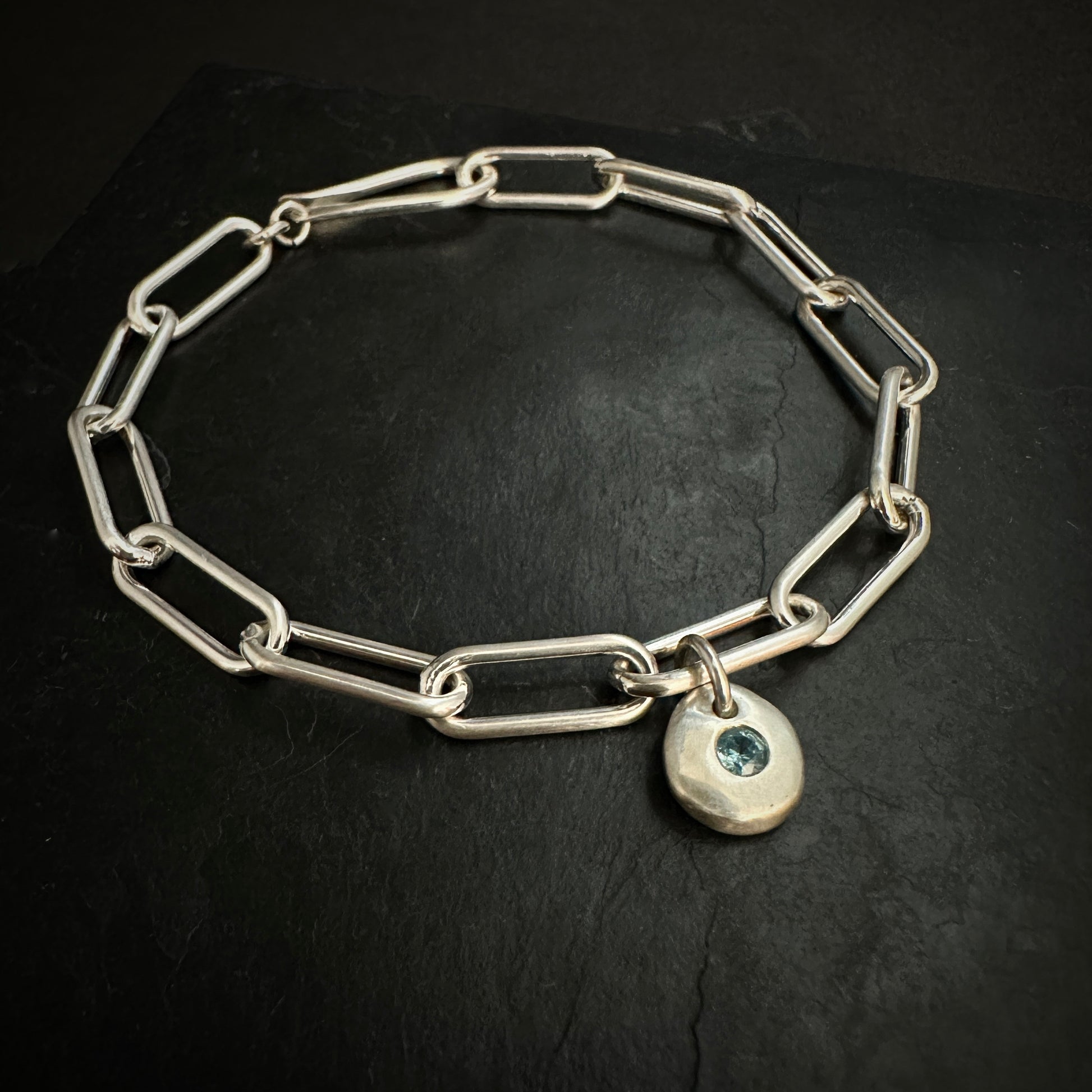 Silver chain bracelet with a heart-shaped charm featuring a blue gemstone on a black background