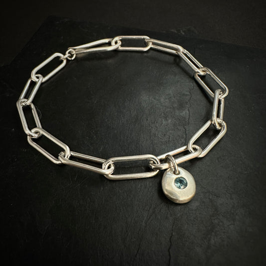 Silver chain bracelet with a heart-shaped charm featuring a blue gemstone on a black background